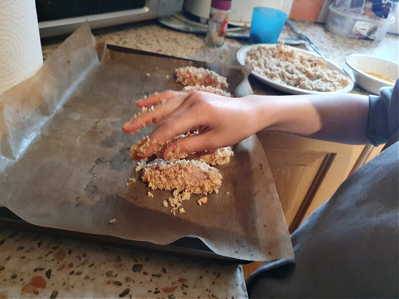 child's hand putting breadcrumbed chicken goujons on a tray