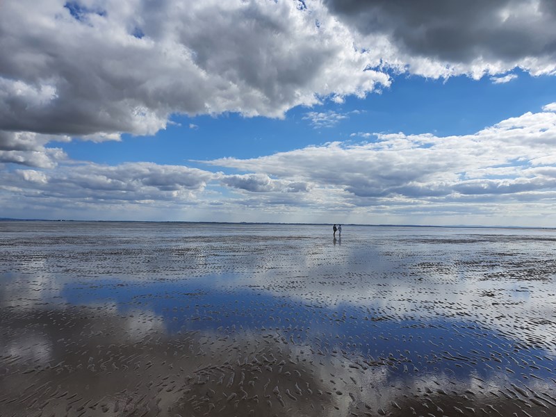 sky and cloud reflections on watery beach with 2 people walking in the distance