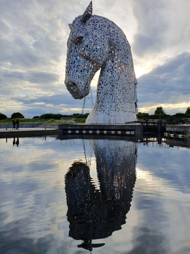 kelpie 1 sculpture with reflection in the water below it and sun and clouds behind it