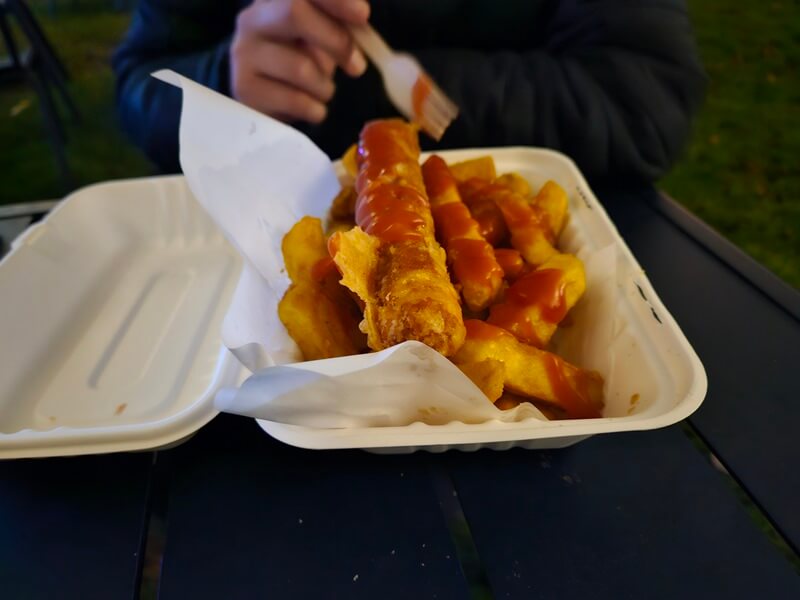 battered sausage and chips in takeaway carton with ketchup over