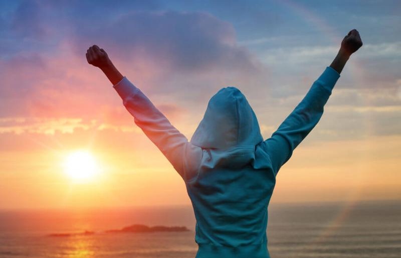 woman with hands in victory pose back to the camera with blue hoodie hood up standing against a sunset